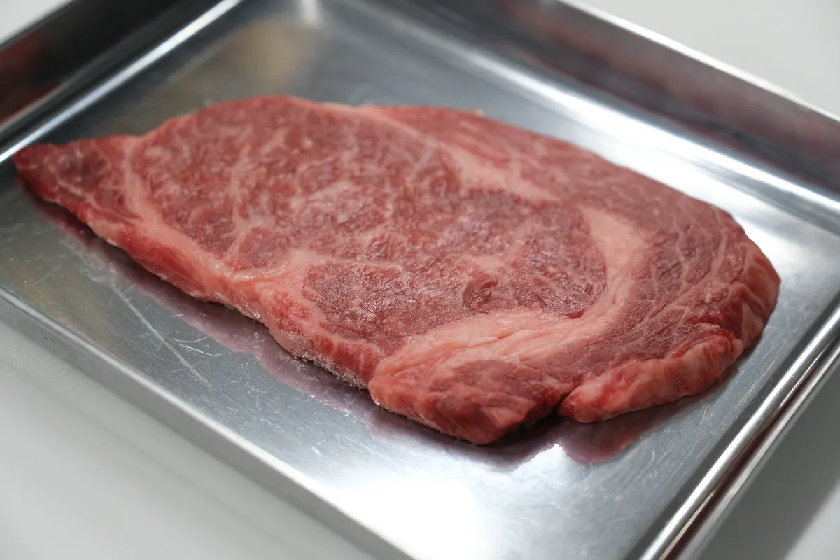 光沢のあるステンレスバットの中央に置かれた解凍後の牛ステーキ肉。赤身に白いサシが網目状に入り、切り口はやや落ち着いた赤みで、表面の照りは控えめ