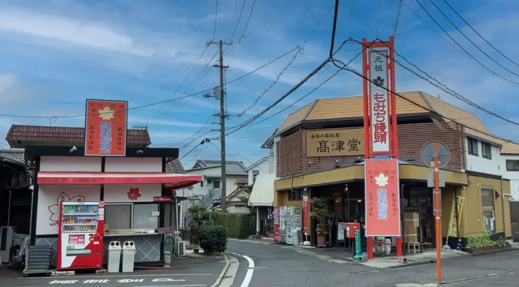 広島・高津堂の外観写真、茶色の建物と赤い看板、元祖もみぢ饅頭の立て看板が店舗前に立つ街角風景