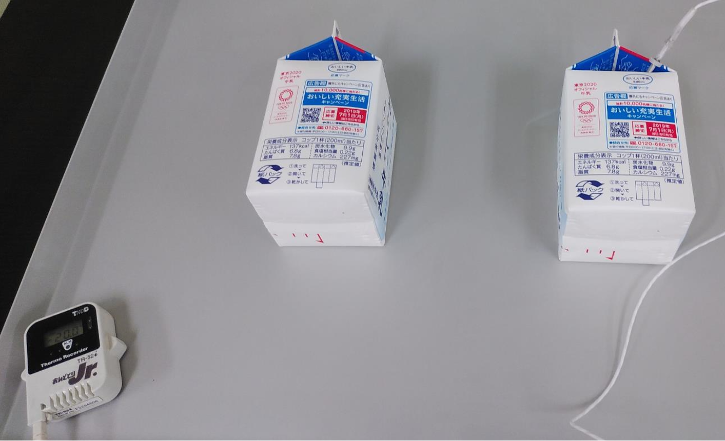 白い牛乳の紙パック2本を立てて置き、右のパック上部に温度センサーのケーブルを挿し込み、手前に置いたデジタル温度記録計で中心温度を計測する凍結前のセッティングの様子