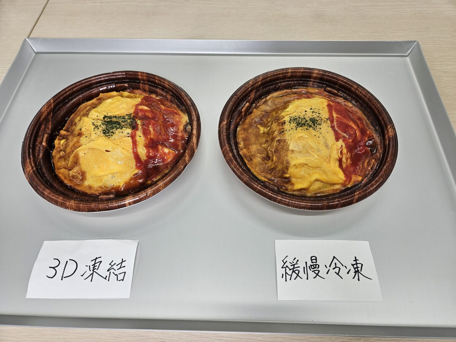 アルミトレー上の茶色い丸容器に入った黄色い卵と赤いソースのオムライスが左右に並び、左に「3D凍結」、右に「緩慢冷凍」の手書き札が置かれている