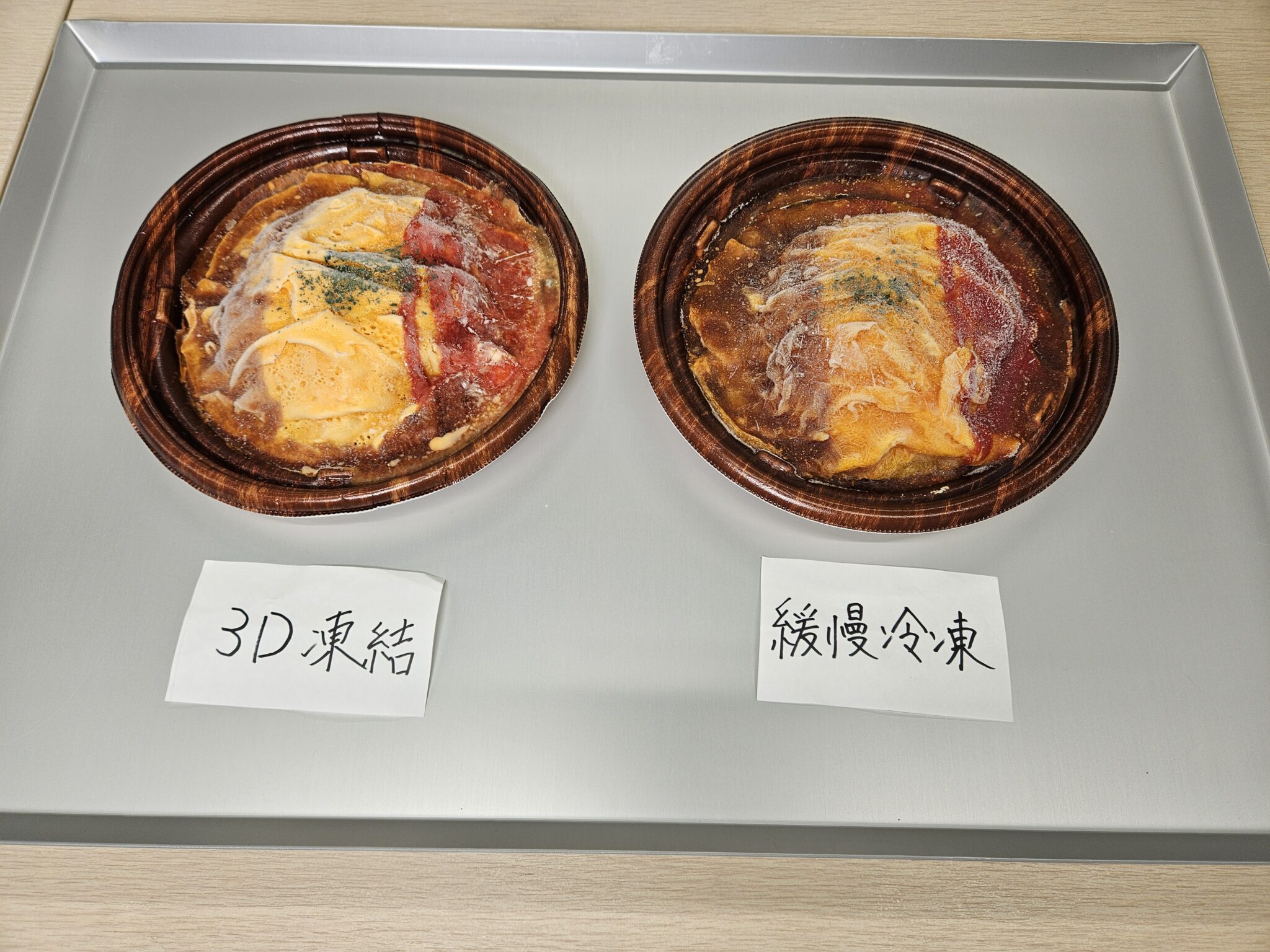アルミトレー上の茶色い丸容器に入ったオムライスが左右に並び、左に「3D凍結」、右に「緩慢冷凍」の手書き札が置かれ、卵と赤いソースの表面に白い霜が見える