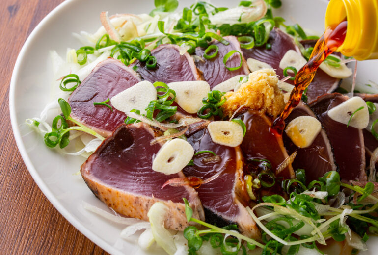 鰹のタタキにポン酢をかける瞬間｜ニンニクスライスやネギ、みょうがが添えられた新鮮なかつお料理