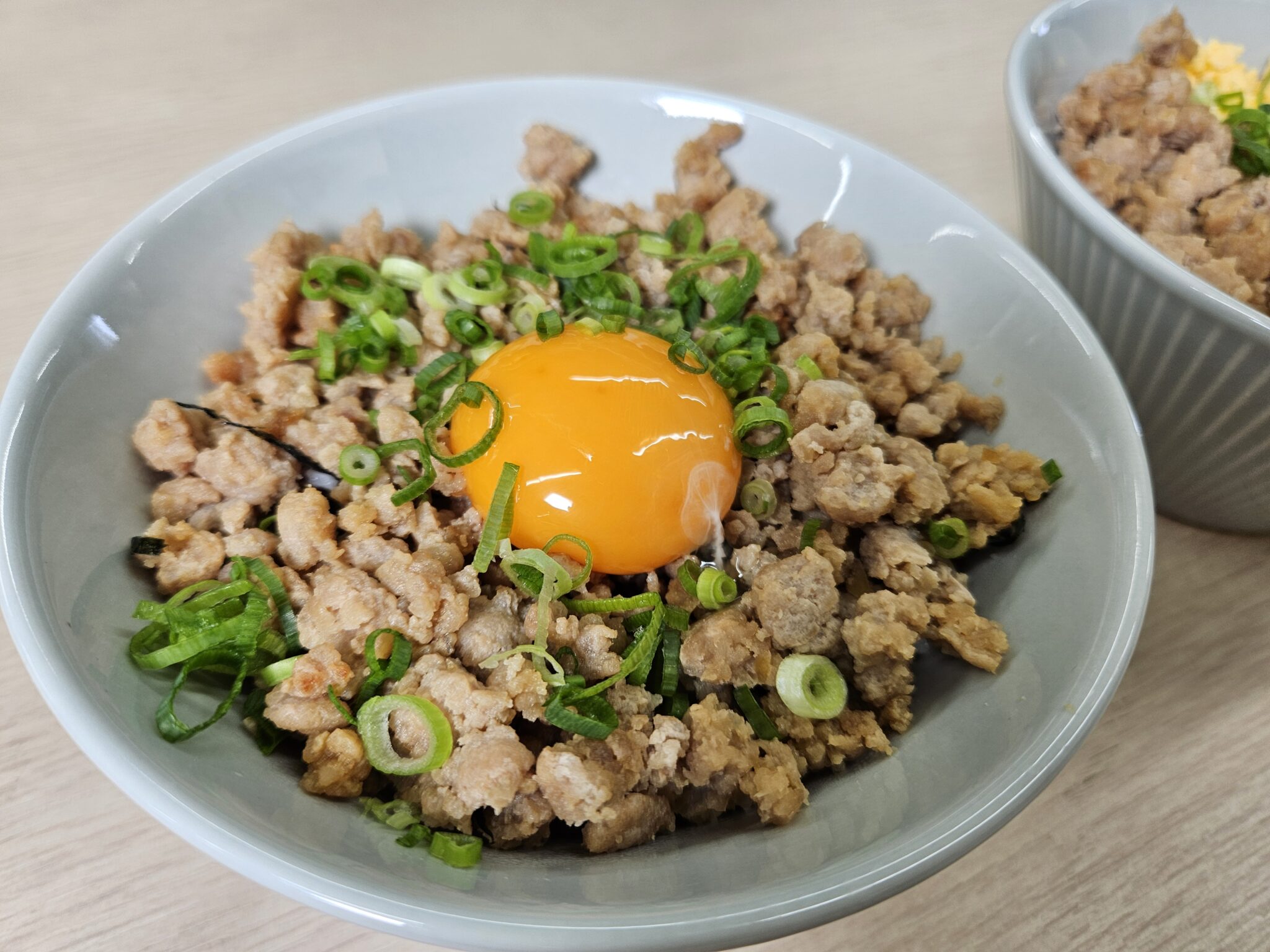 灰色の丼に盛った茶色い鶏そぼろの中央に、つやのある卵黄をのせ、小口切りの青ねぎを散らした鶏そぼろ丼。奥にもう一杯の丼が写る。