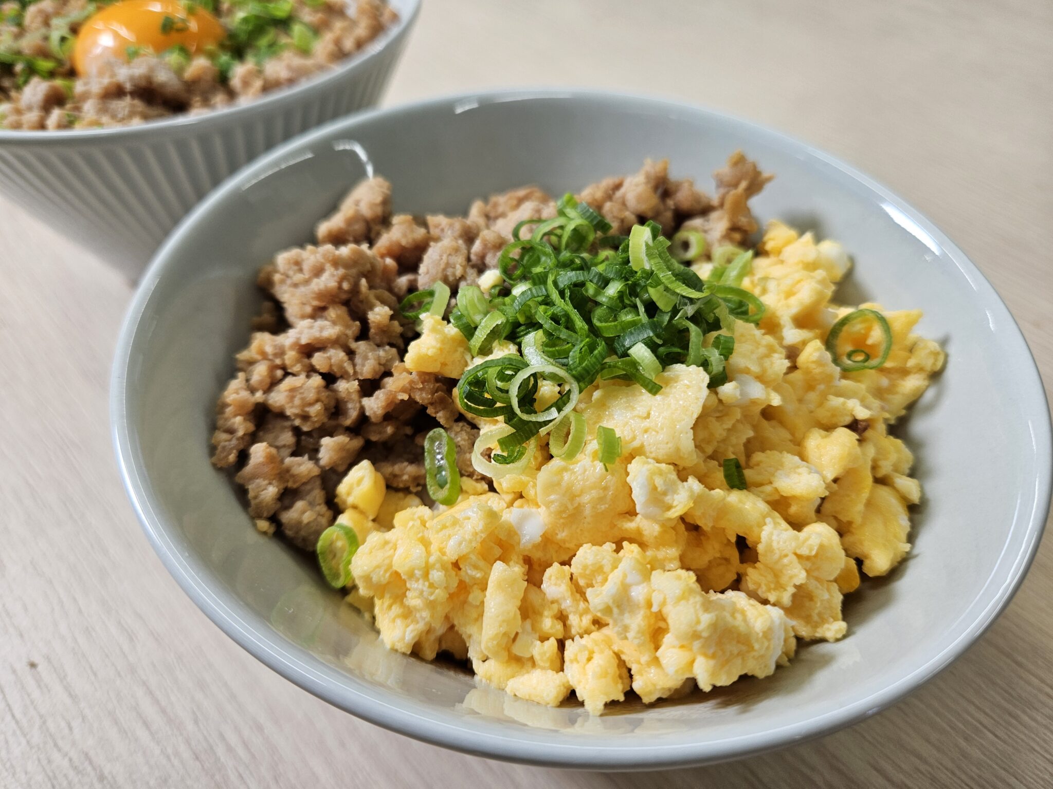 灰色の丼に、茶色い鶏そぼろとふんわりした黄色い炒り卵を左右に盛り分け、中央に小口切りの青ねぎをのせた二色丼。奥にもう一杯が写る。