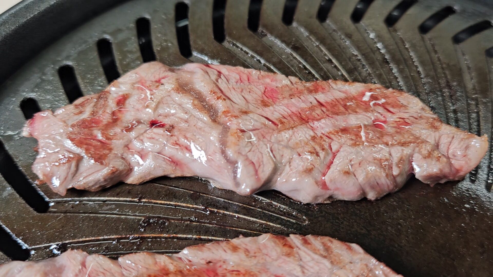 黒い焼肉プレートの上で焼かれている横長の牛肉の接写。淡い桃色の表面に細い赤色の筋と茶色い焼き目があり、周囲に光を反射する液体が見える