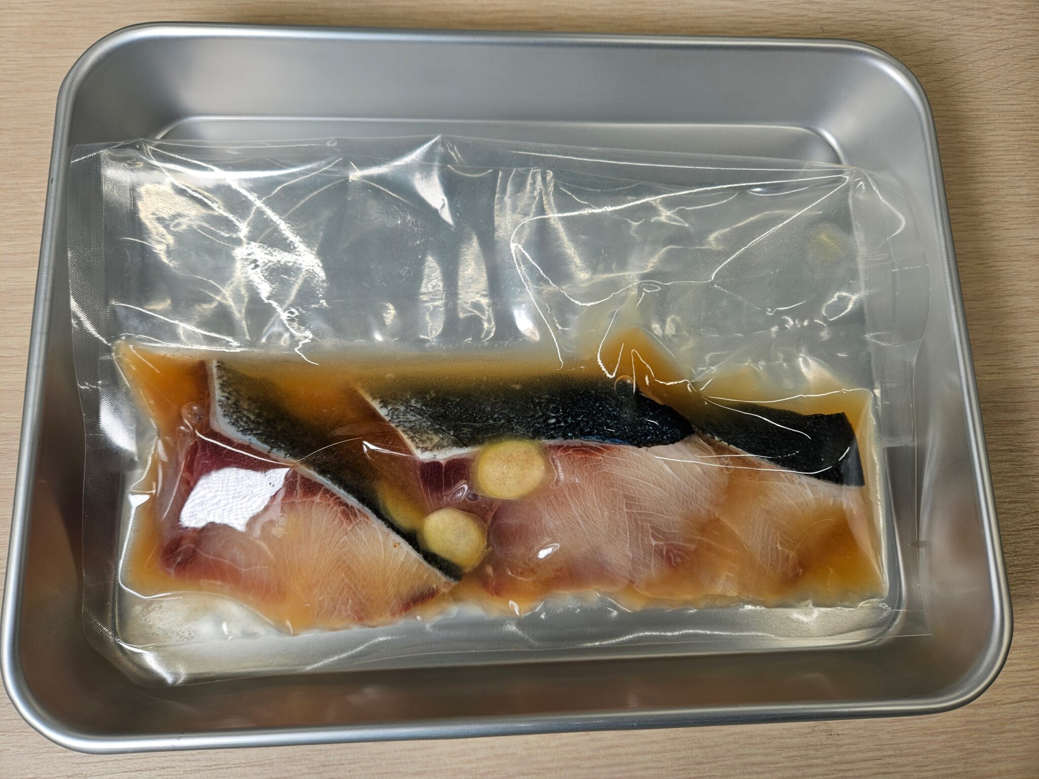透明な袋に入れたタレ漬けのブリの切り身。琥珀色の調味液と皮・血合いのある身が見え、アルミトレーにのっている