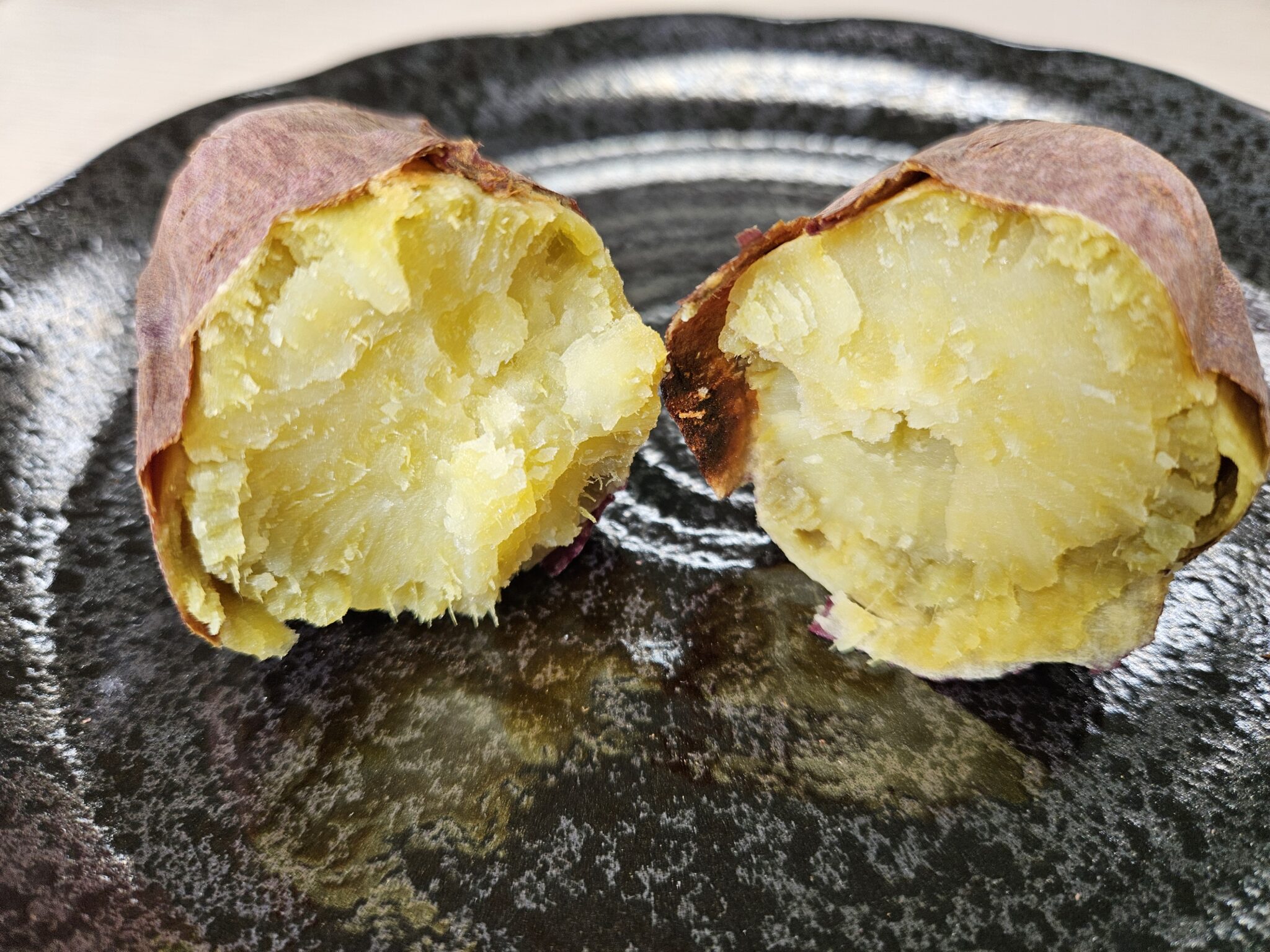 焼き芋を横に割った断面2つのクローズアップ。黄色くしっとりした果肉と紫色の皮、黒い皿につやのある解凍後の断面