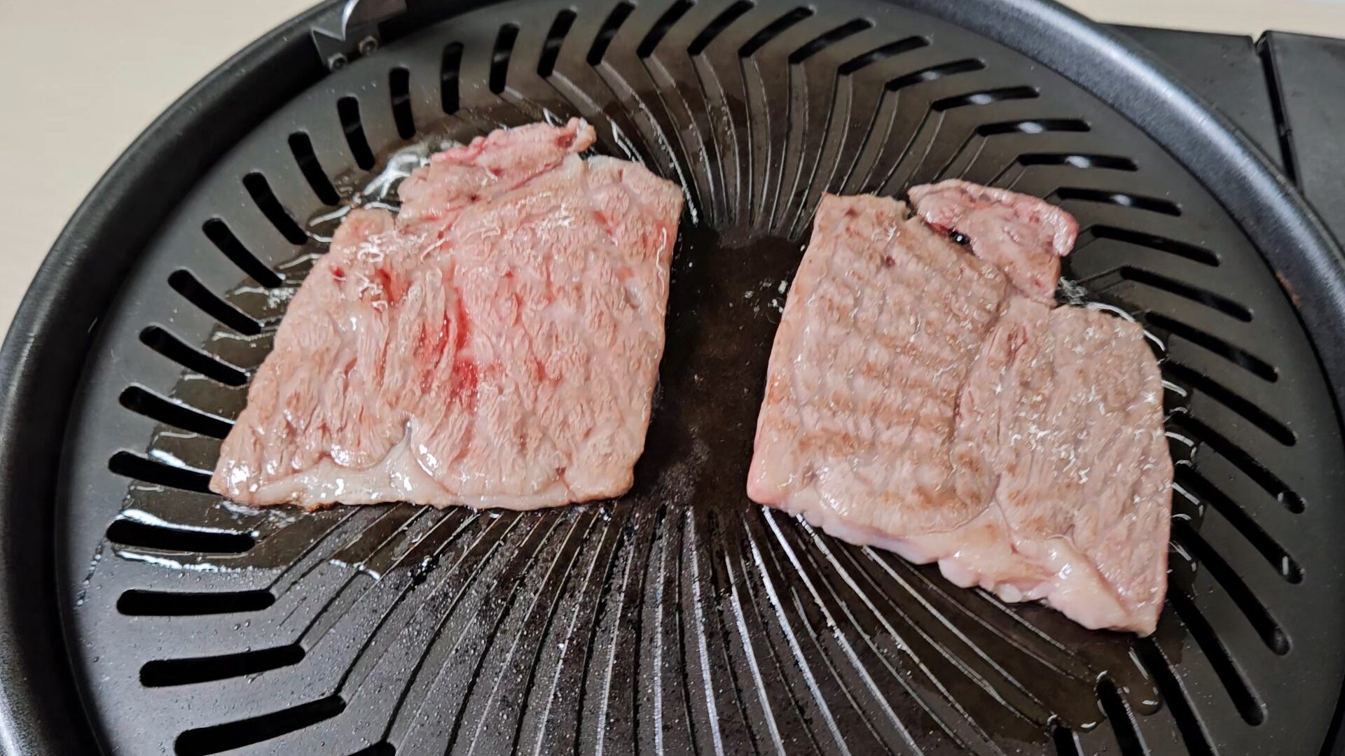 放射状の溝がある黒い円形の焼肉プレートに、淡い桃色へ焼けた薄い牛肉を左右に置き、肉の周囲へ透明な液体が広がっている様子