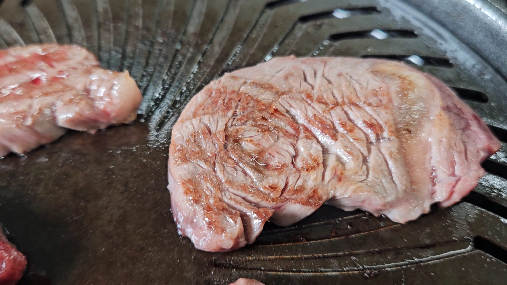黒い焼肉プレートの上で焼かれている厚みのある楕円形の牛肉の接写。淡い桃色の表面に渦状の筋と茶色い焼き目が入り、奥にも別の肉が見える