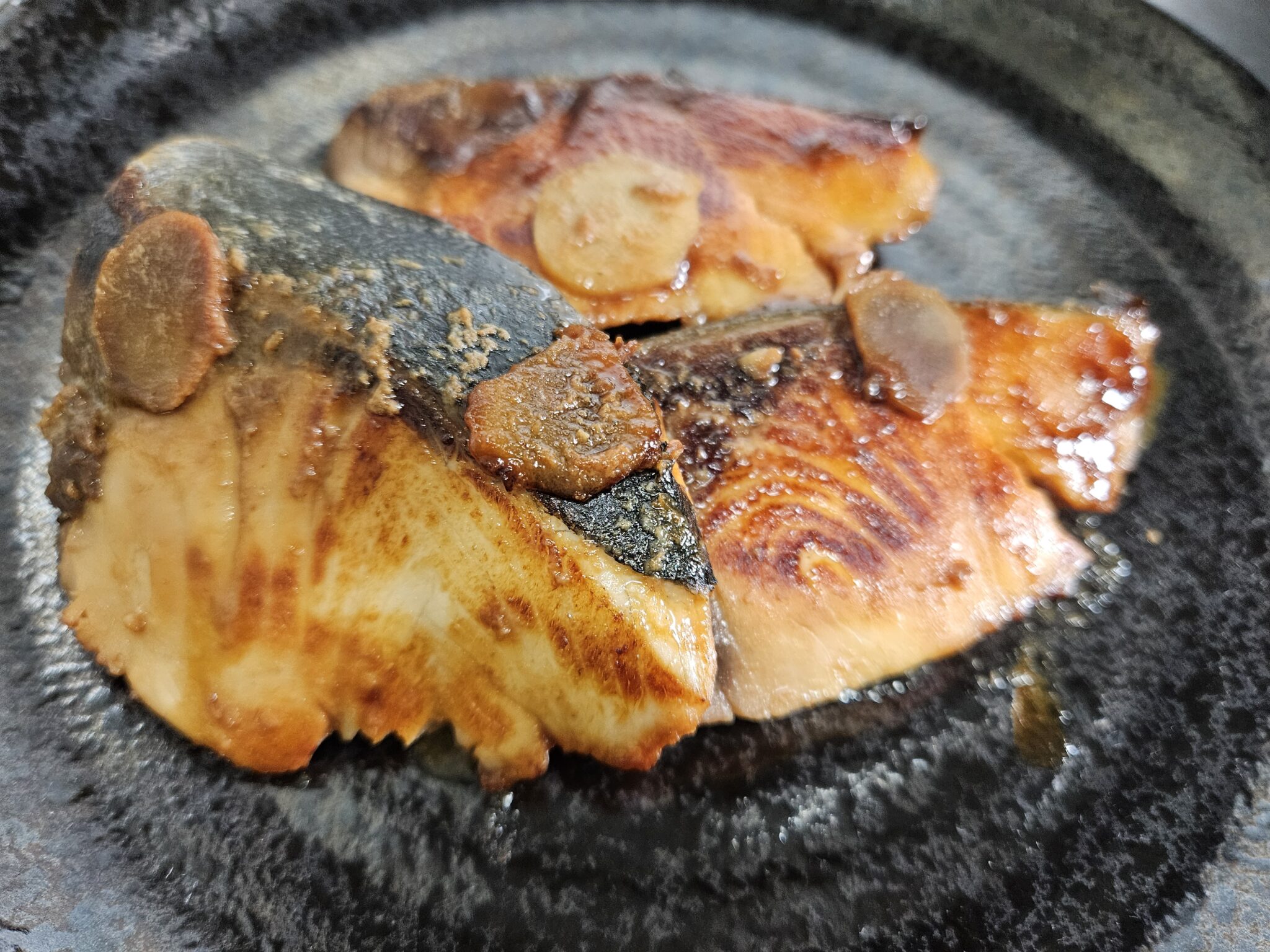 黒い器に盛った焼き上がりのブリの照り焼き。ツヤのあるタレと焼き目のついた身、皮、しょうがのスライスが見える