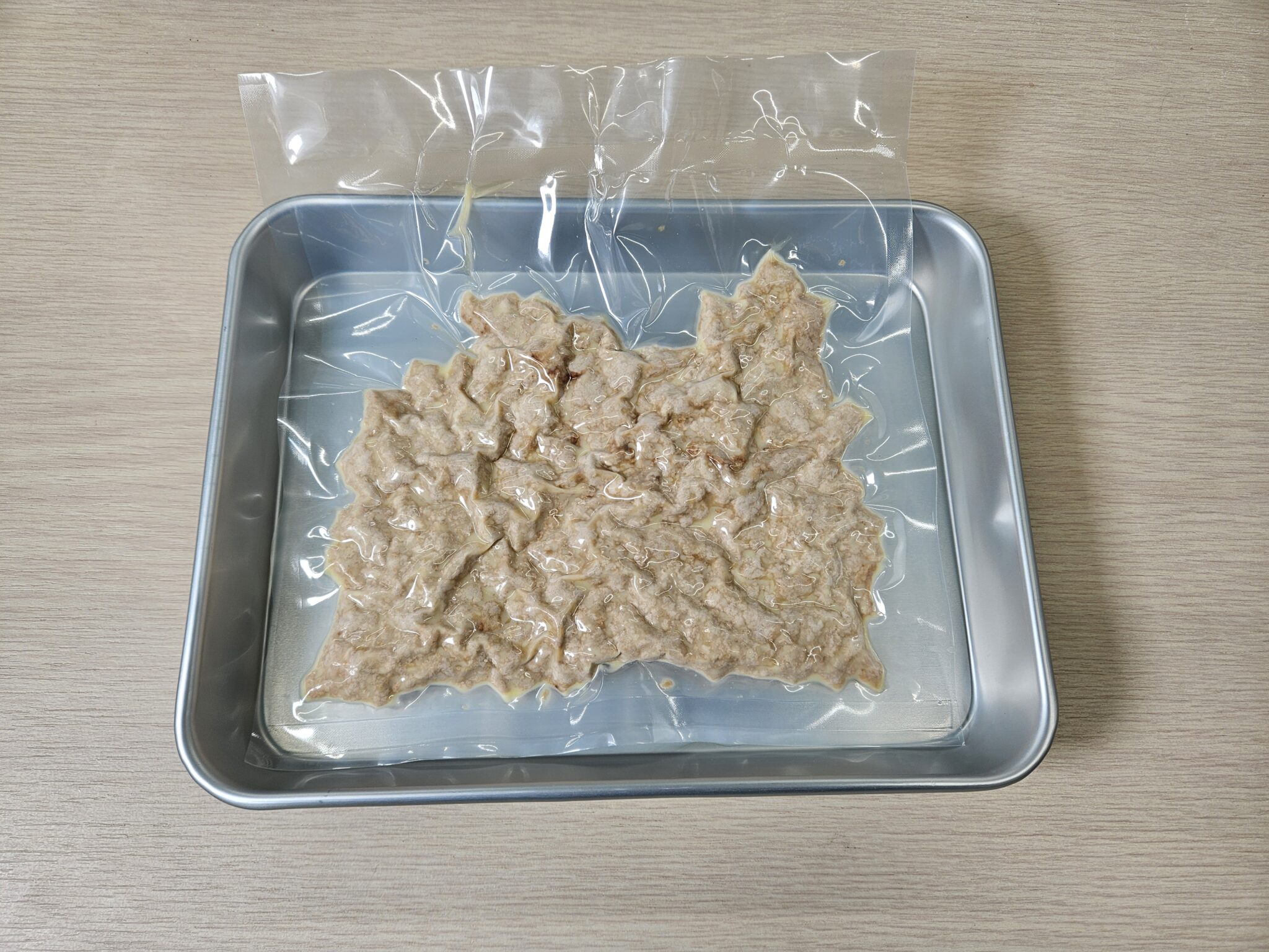 真空パックにして平らに広げた鶏そぼろを、青みがかったアルミトレーに乗せた凍結前のサンプル。木目のテーブルの上に置かれ、袋の中でミンチが均一な厚みに広がっている。