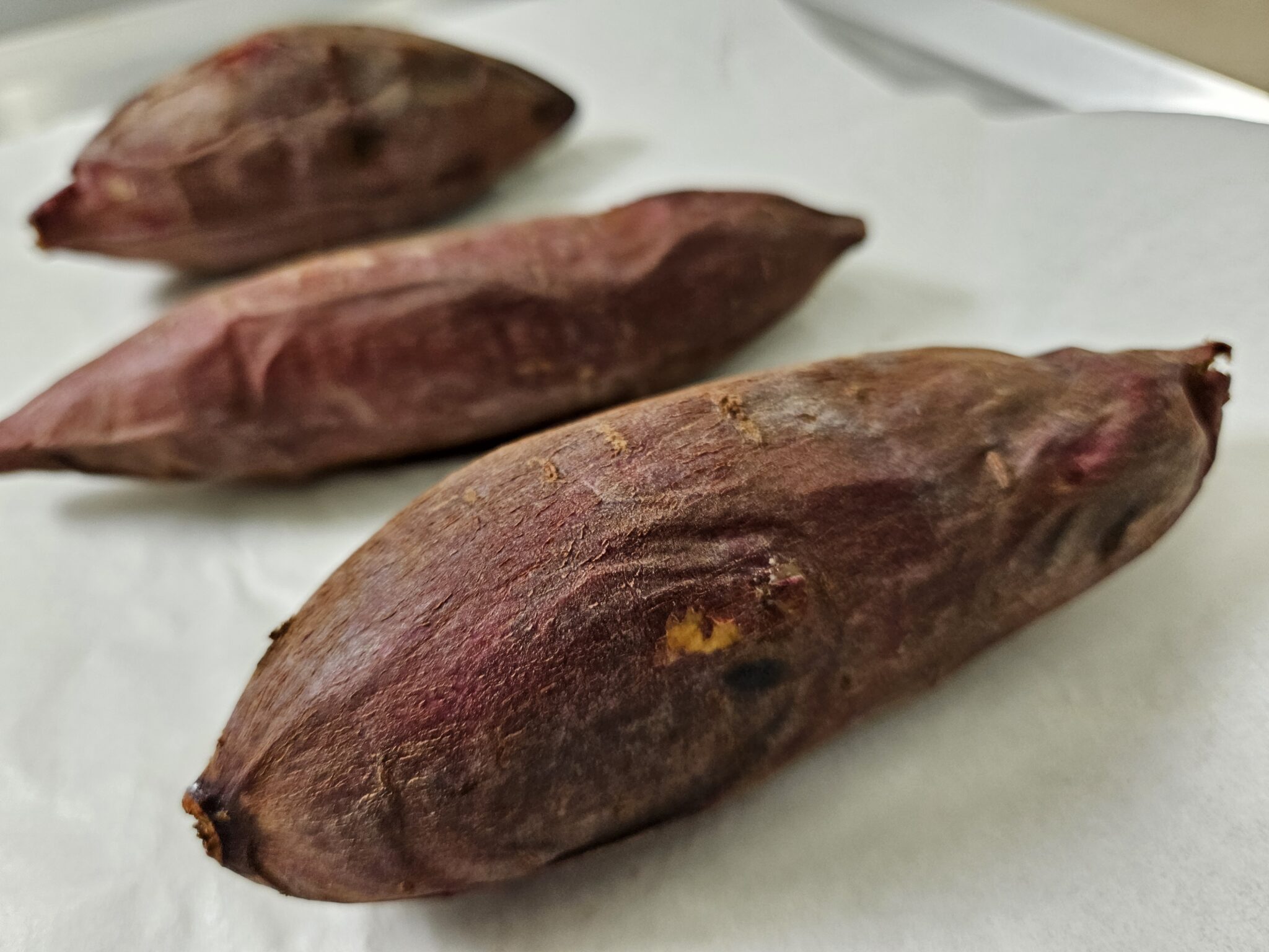 白いトレーに並べた赤紫色の皮の焼き芋3本のクローズアップ。皮は乾いてマットな質感で、ややシワが寄った焼き上がりの表面