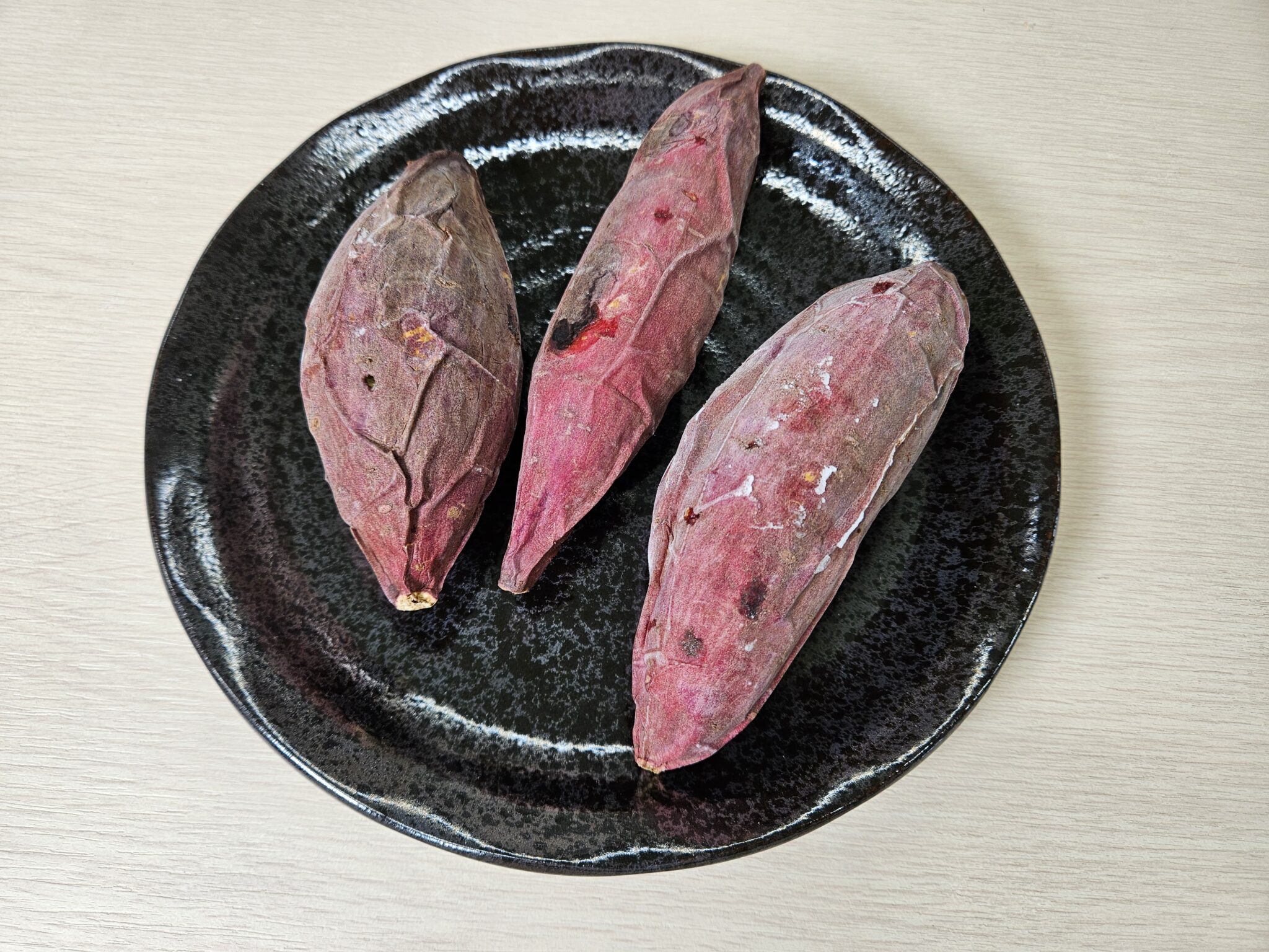 黒い丸皿に並ぶ赤紫色の皮の焼き芋3本。表面に薄く白い霜がのり、ややシワの寄った皮のまま丸ごとの形を保った急速冷凍後の仕上がり
