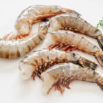 殻をむいたブラックタイガー無頭エビを並べた写真 白背景に緑の葉を添えた新鮮な冷凍用食材