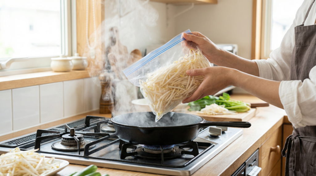 日本人女性が冷凍保存袋から凍ったままのもやしを取り出し、湯気が立つ熱いフライパンに投入している様子。明るい日本の家庭のキッチン