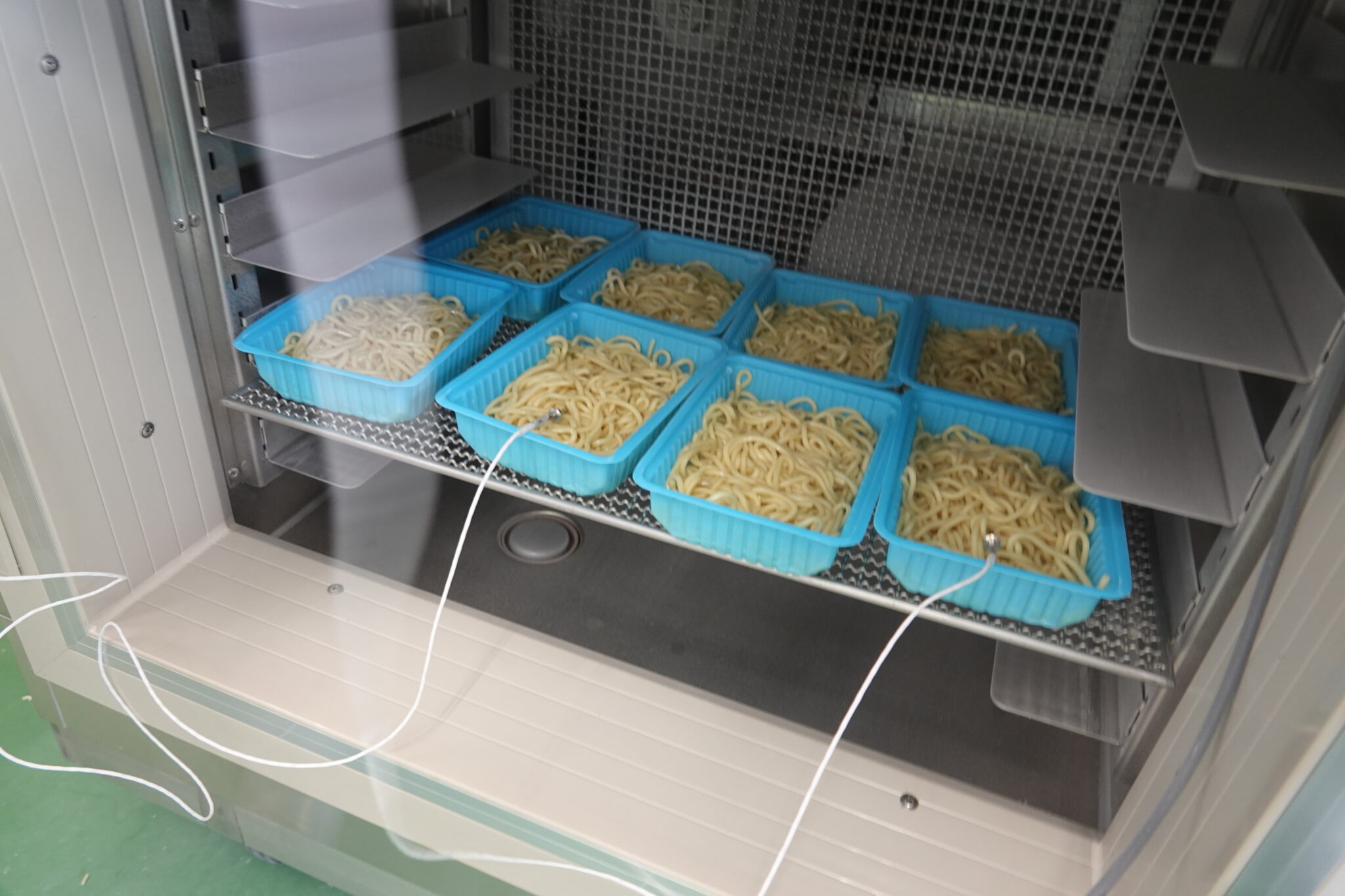 開いた金属製庫内の棚に、太い麺を入れた青いトレーを複数並べ、手前のトレー2枚へ白いコードの温度センサーを差し込んだ様子