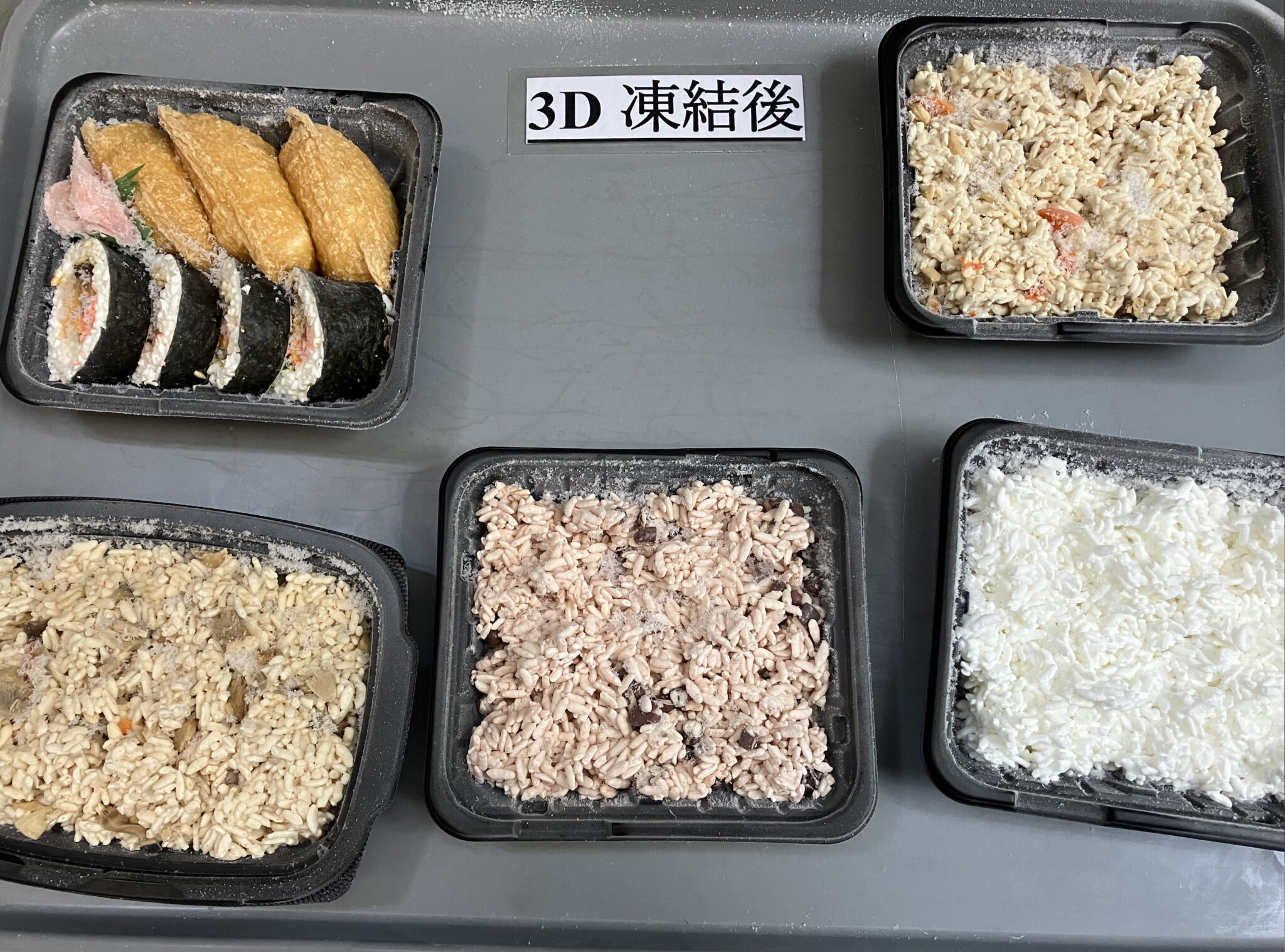 トレイ上の黒い容器に凍結した白米、赤飯、鶏飯、炊き込みご飯、いなり寿司と海苔巻きを並べ、中央に「3D凍結後」と表示した写真