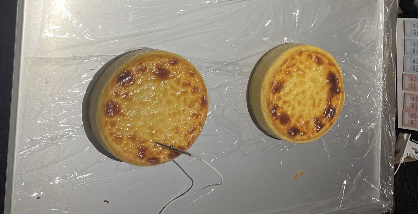 ラップを敷いたステンレス台に大きさの異なる焼き色付きの丸いチーズケーキを2台置き、左のホール中央付近へ温度プローブを差した様子